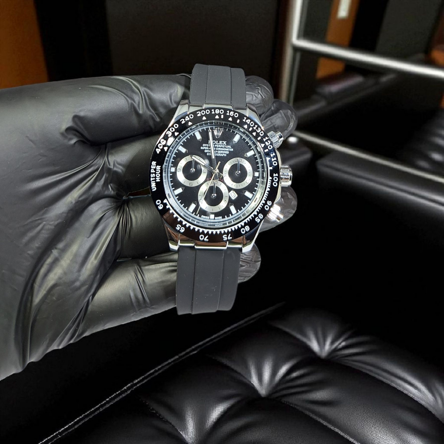 Luxury watch with black dial and gray strap held by a gloved hand on a black leather surface.